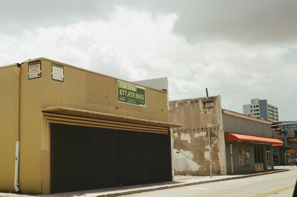 Photograph of a street with buildings that appear old and abandoned. A for sale sign is visible on one of the buildings. 